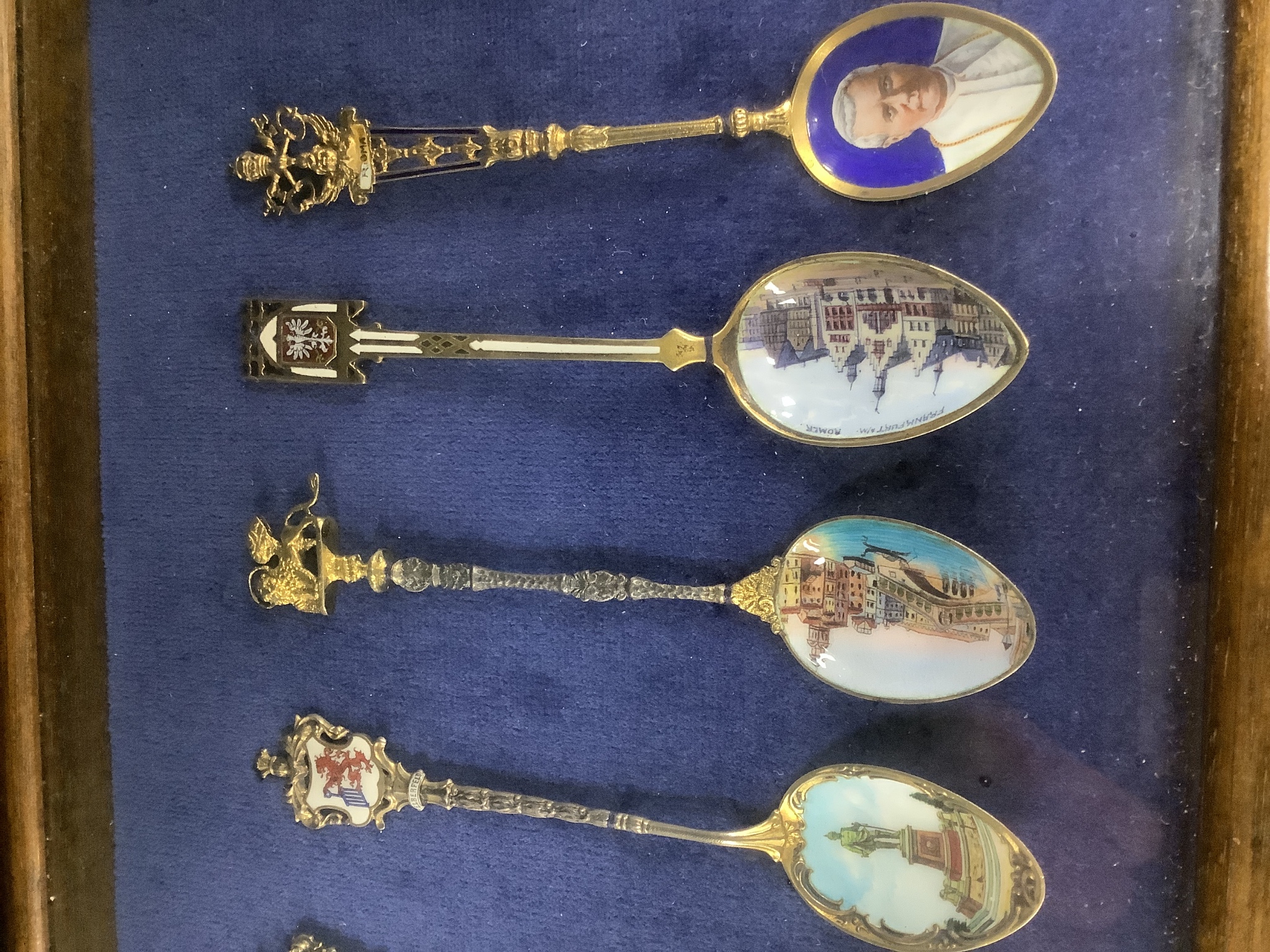 A collection of sixty four 800 standard silver (many gilt) and enamel souvenir spoons, early 20th century, in eight display cases, Including views of London, Germany, Italy, Austria, Spain etc.